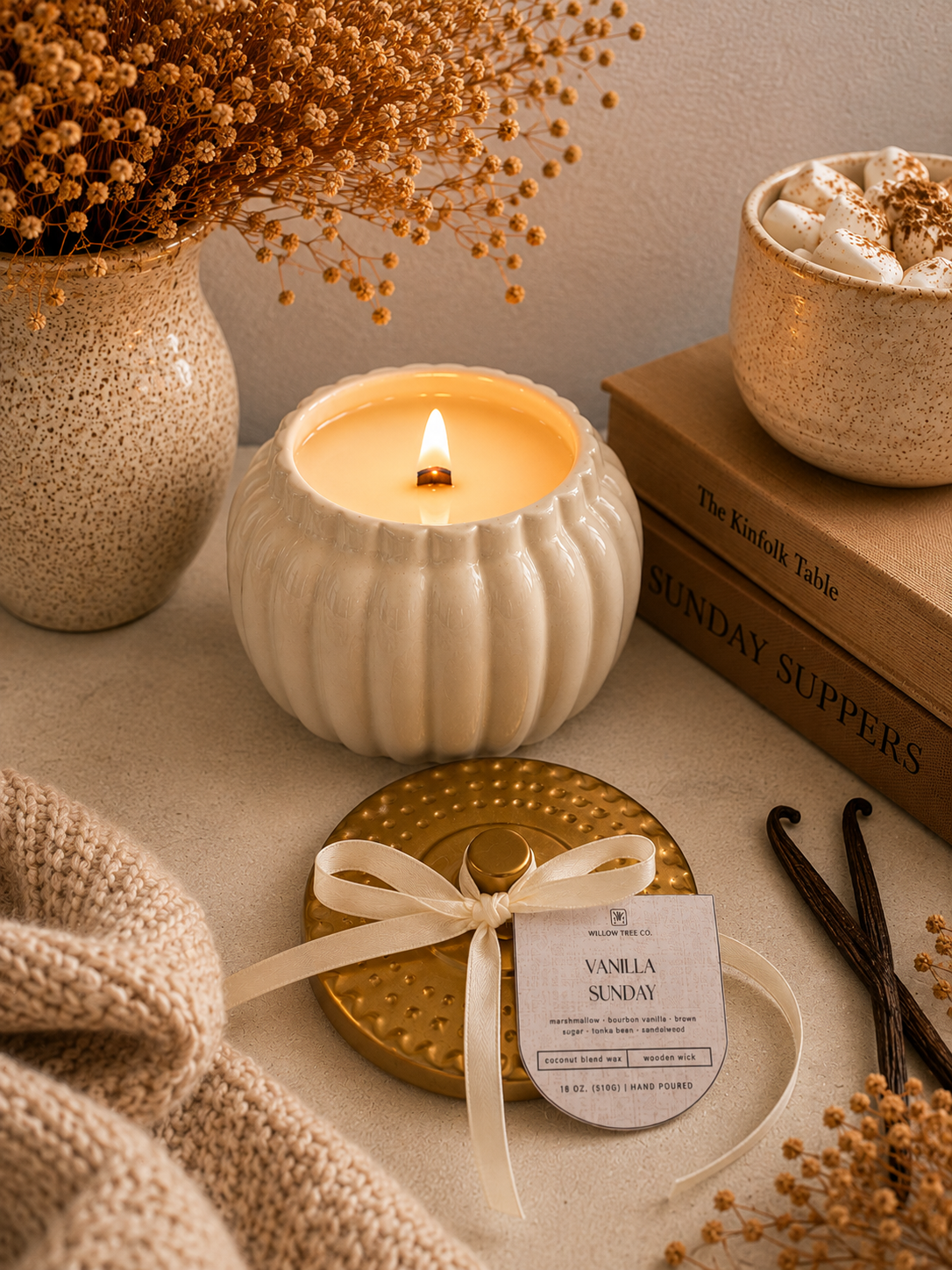 A cozy tabletop scene featuring a lit cream-colored ribbed candle holder with a small flame, alongside a gold-toned lid tied with a cream ribbon and a tag reading 'VANILLA SUNDAY.' Decorative props include dried flowers in a speckled vase, stacked books, a knit blanket, vanilla beans, and a bowl of whipped cream.