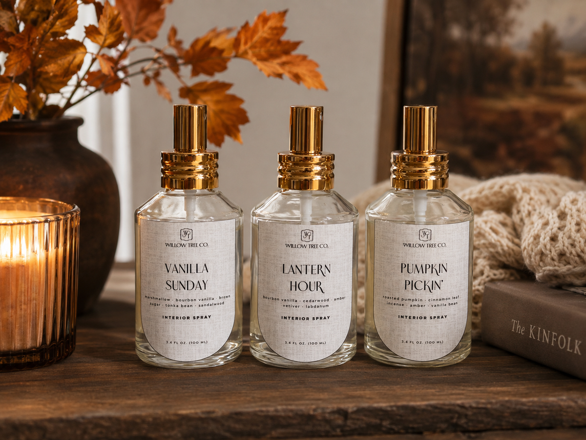 Three clear glass perfume bottles with gold caps and white labels are arranged on a wooden surface. The labels read Vanilla Sunday, Lantern Hour, and Pumpkin Pie.
