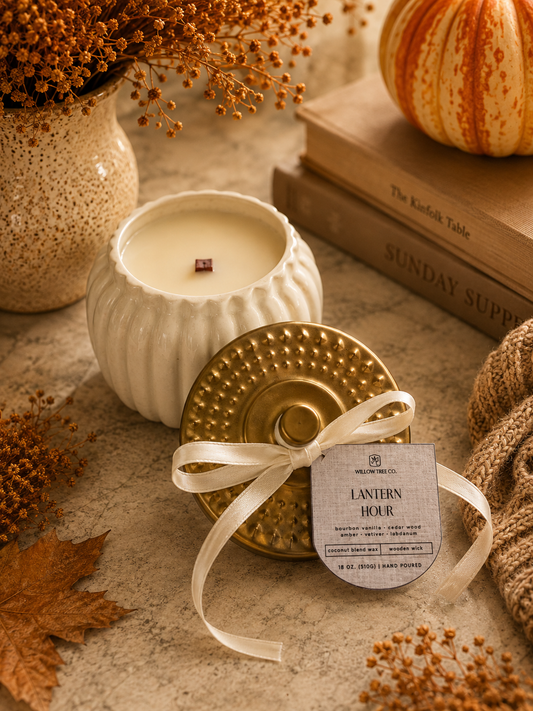 A styled product photo of a candle in a ribbed white ceramic jar with its lid placed in front. The lid is metallic gold with a white ribbon tied around it and a paper tag that reads 'LANTERN HOUR.