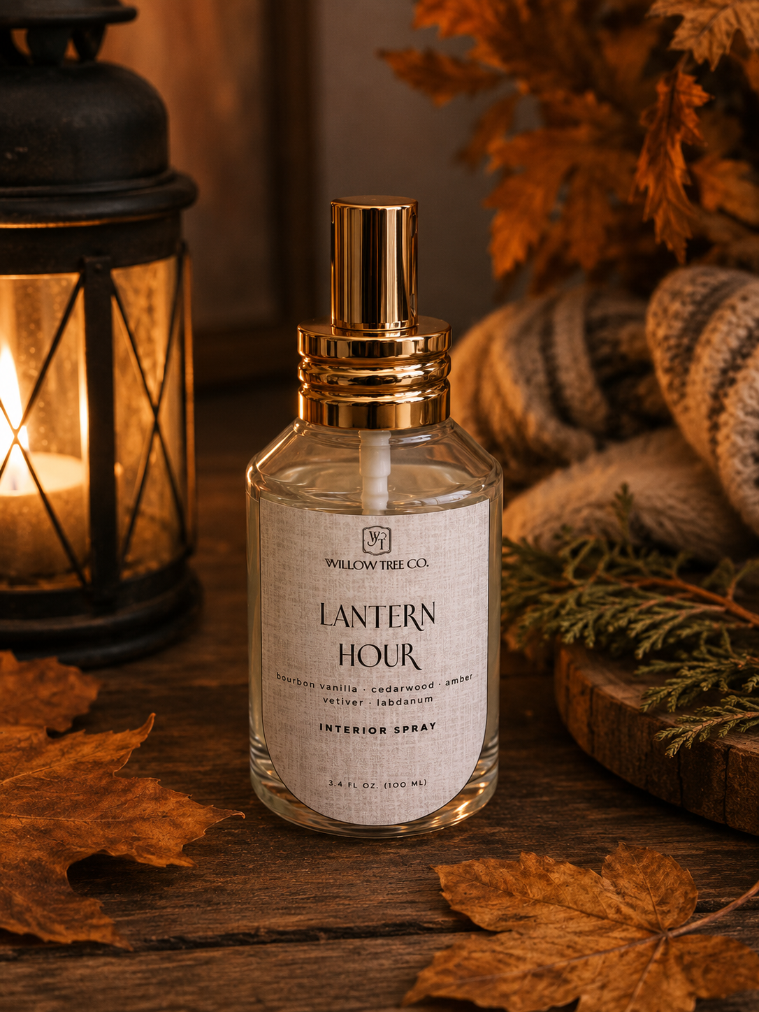 A clear glass spray bottle labeled 'Lantern Hour' by Willow Tree Co., with a gold atomizer cap, styled on a rustic wooden surface with autumn leaves, a lit lantern, and cozy knit textiles in the background.