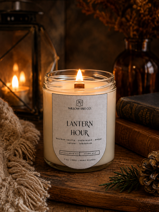 A lit candle in a clear glass jar sits on a rustic wooden surface with a label reading 'LANTERN HOUR' and 'WILLOW TREE CO.' Warm candles, books, a lantern, and dried flowers create a cozy styled scene.
