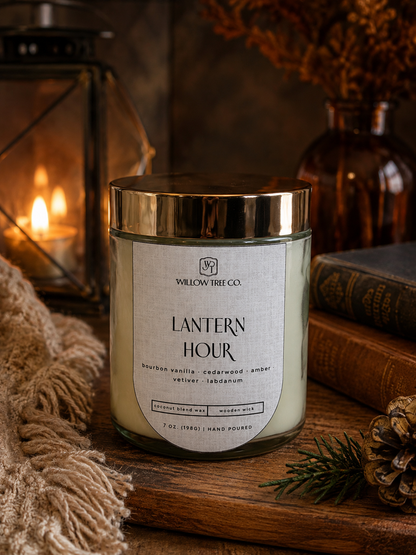 A candle in a clear glass jar with a shiny gold lid and a cream label reading 'LANTERN HOUR' by Willow Tree Co., styled on a wooden surface with a lantern, books, pinecone, and dried flowers in the background.