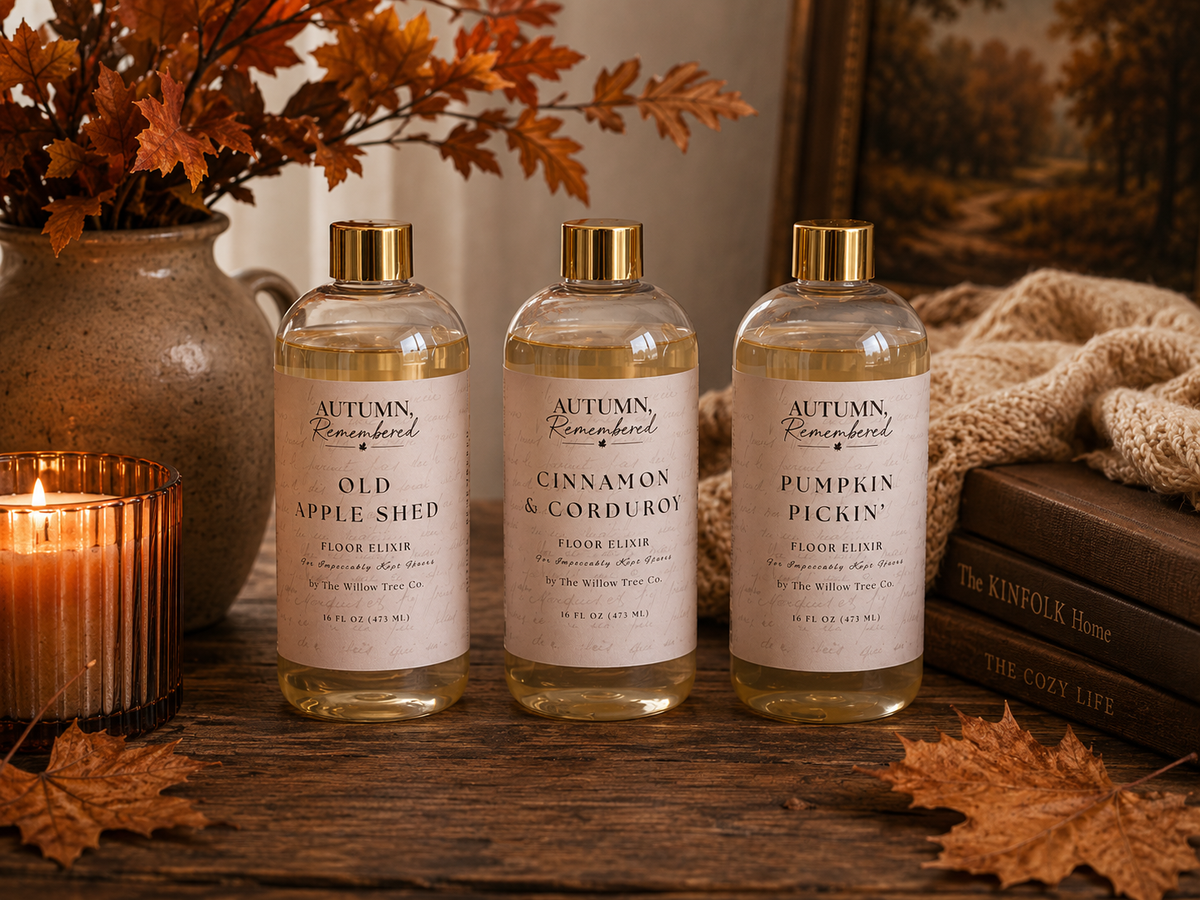 Three clear bottles of Autumn, Remembered floor elixirs labeled Old Apple Shed, Cinnamon & Corduroy, and Pumpkin Pickin', arranged on a rustic wooden table with autumn-themed props including a candle, books, and dried leaves.