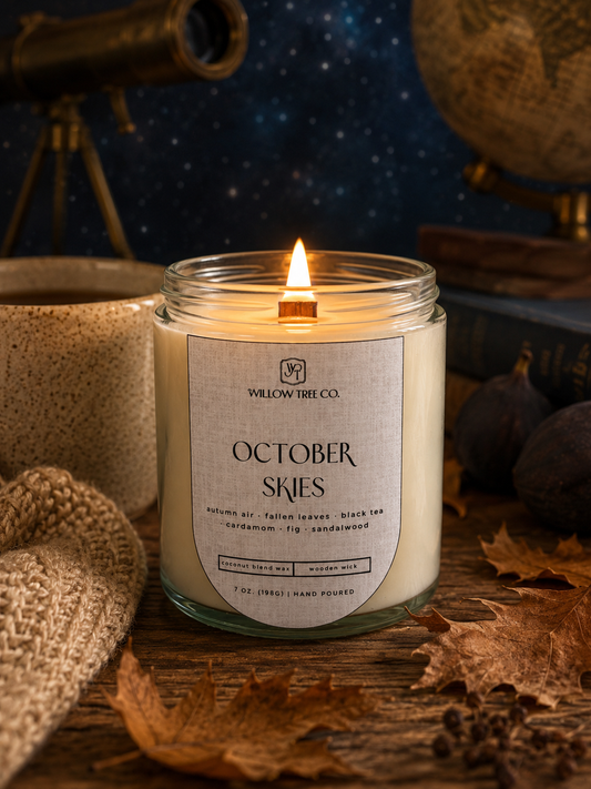 A lit candle in a clear glass jar with a cream-colored wax and a white label reading 'WILLOW WREE CO. OCTOBER SKIES' on a rustic wooden surface surrounded by autumn leaves, a knit blanket, and a globe in the background.