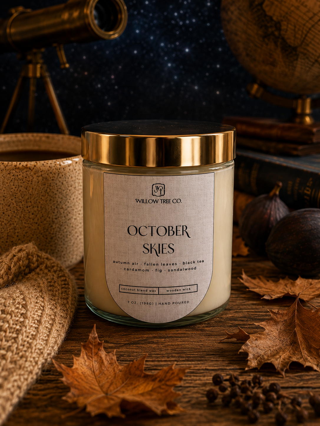 A candle jar labeled 'October Skies' by Willow Tree Co., styled on a rustic wooden surface with autumn leaves, a knit sweater, books, and a globe in the background.