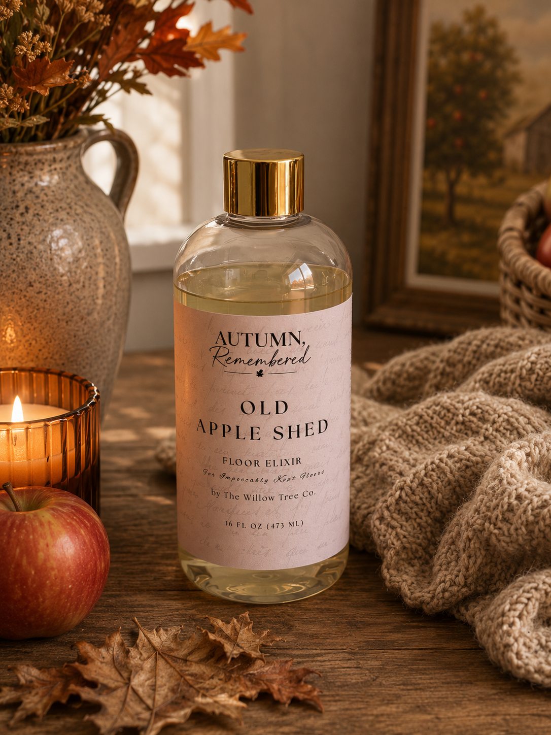 A bottle of Autumn Remembered Old Apple Shed floor elixir sits on a rustic wooden table surrounded by cozy fall-themed props, including a lit candle, a red apple, dried leaves, a knitted blanket, and a ceramic vase with autumn foliage.