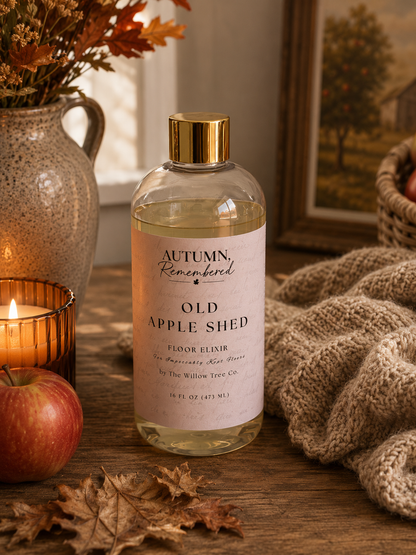 A bottle of Autumn Remembered Old Apple Shed floor elixir sits on a rustic wooden table surrounded by cozy fall-themed props, including a lit candle, a red apple, dried leaves, a knitted blanket, and a ceramic vase with autumn foliage.