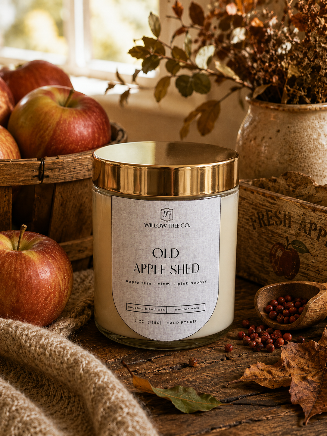 A candle jar labeled 'Old Apple Shed' by Willow Tree Co., styled on a rustic wooden surface with apples, dried leaves, a burlap cloth, and a ceramic vase in the background.