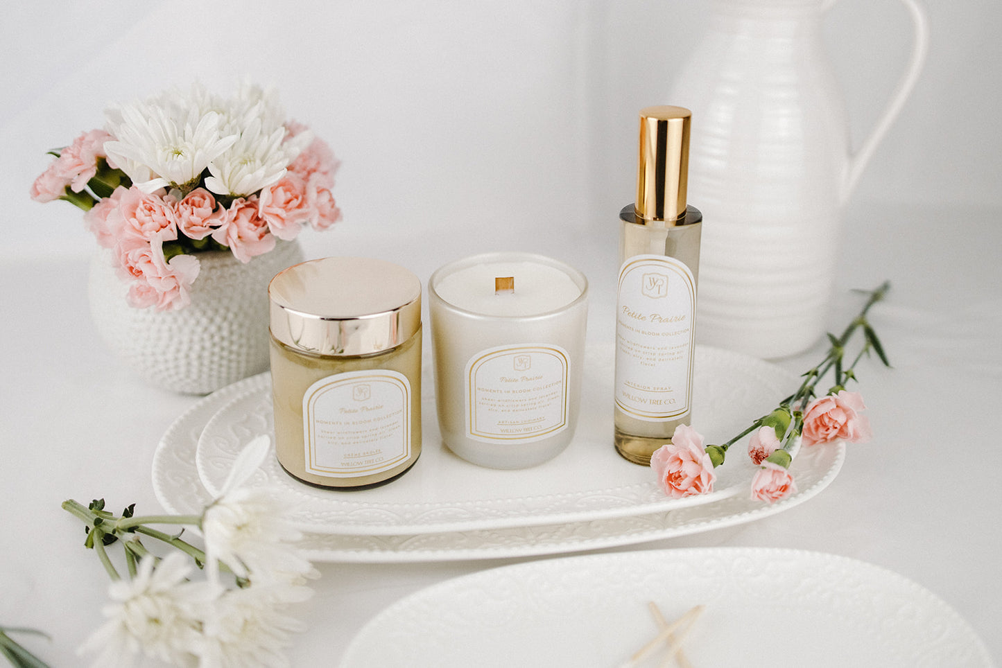 Candle, body cream, and interior spray set on a white tray with flowers in the background