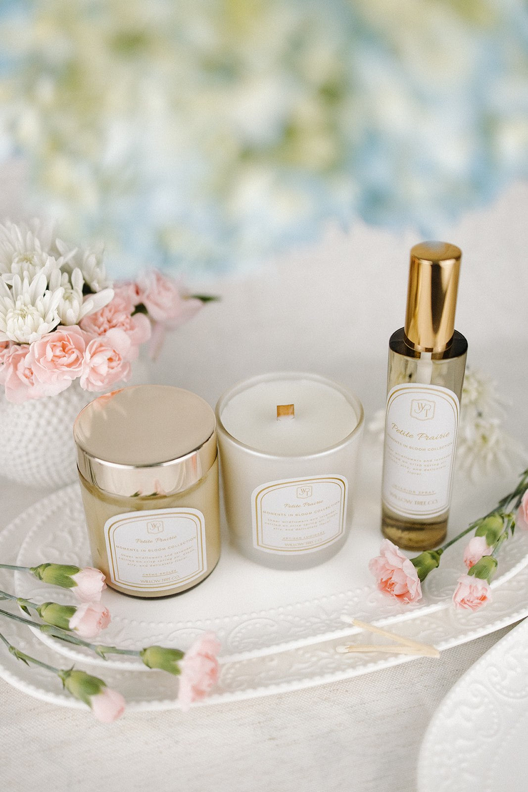 Interior spray, candle, and a jar of body cream on a white surface with flowers in the background