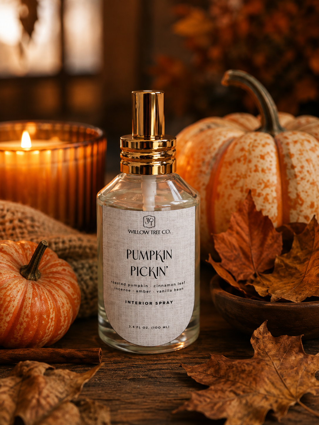 A clear glass interior spray bottle labeled 'Pumpkin Pickin'' by Willow Tree Co., with a gold atomizer cap, styled on a wooden surface with autumn leaves, small pumpkins, burlap, and a lit candle in the background.