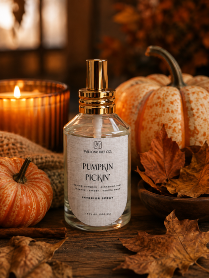 A clear glass interior spray bottle labeled 'Pumpkin Pickin'' by Willow Tree Co., with a gold atomizer cap, styled on a wooden surface with autumn leaves, small pumpkins, burlap, and a lit candle in the background.
