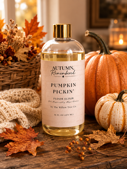 A clear bottle of Autumn, Remembered Pumpkin Pickin' Floor Elixir by The Willow Tree Co. sits on a rustic wooden table surrounded by autumn decorations including pumpkins, fall leaves, and a knitted blanket.