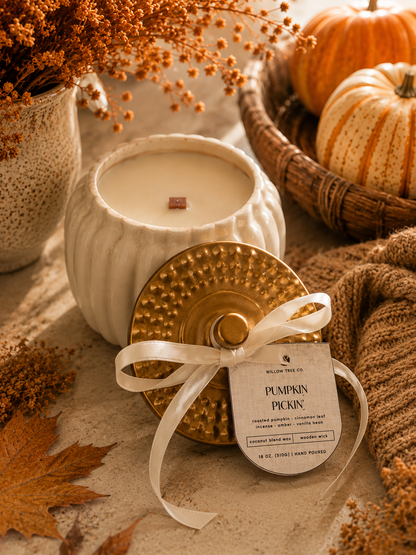 A styled product photo of a 'Pumpkin Pickin' scented candle by Willow Tree Co. The candle is shown in a white ribbed ceramic jar alongside its gold textured lid, which is tied with a white satin ribbon and a paper tag.