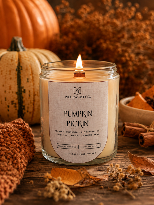 A lit candle in a clear glass jar labeled 'Pumpkin Pickin'' by Willow Tree Co., styled on a wooden surface with pumpkins, cinnamon sticks, dried leaves, and autumn decor in the background.