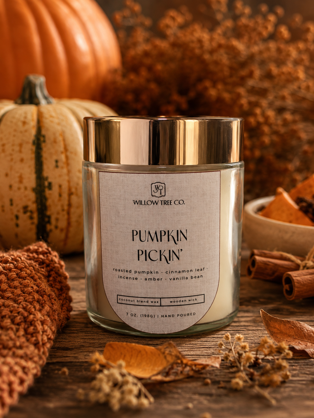 A glass jar candle labeled 'Pumpkin Pickin'' by Willow Tree Co., styled on a rustic wooden surface with pumpkins, cinnamon sticks, dried leaves, and a knitted textile in warm autumn tones.