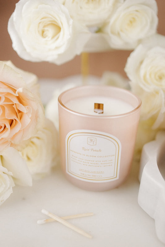 Soft pink rose petals and warm light create a romantic spring setting inspired by Rosé Petale luxury candle and home fragrance, Blush pink candle with a label surrounded by white and peach roses on a marble surface.