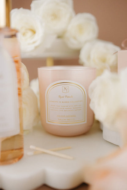 Pink candle with a gold label surrounded by white flowers on a beige surface