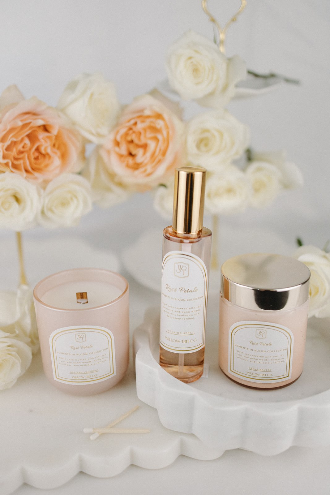 Three luxury products including a candle, interior spray, and jar of body cream on a white surface with flowers in the background.