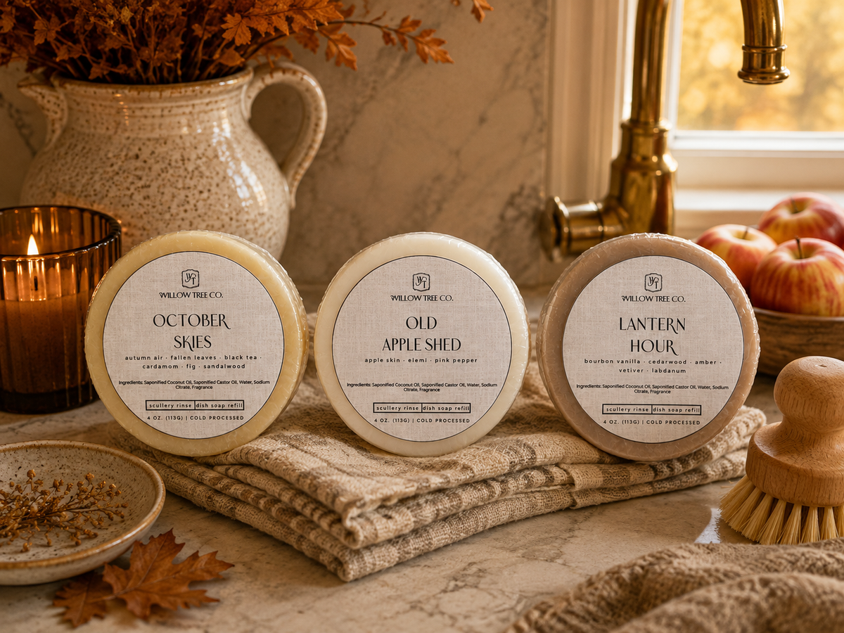 Three round scented solid dish soap bars from Willow Tree Co. labeled 'October Skies', 'Old Apple Shed', and 'Lantern Hour' arranged on a textured beige towel on a kitchen counter, surrounded by fall-themed props including apples, a candle, a vase, and a wooden brush.