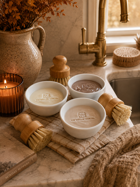 Three small white bowls containing creamy soap bars in off-white, tan, and brown tones, arranged on a marble countertop near a sink with wooden brush holders, a candle, and dried flowers.