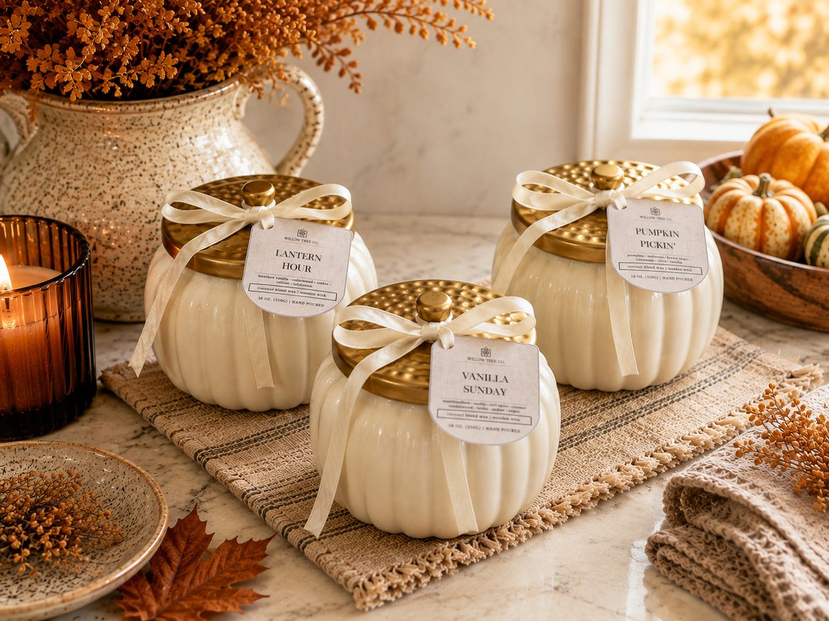 Three decorative pumpkin-shaped candles in cream-colored jars with gold lids and white ribbon bows, labeled 'Pumpkin Pickin' and 'Vanilla Sunday,' styled on a woven mat with fall decor including pumpkins, a candle, and dried leaves.