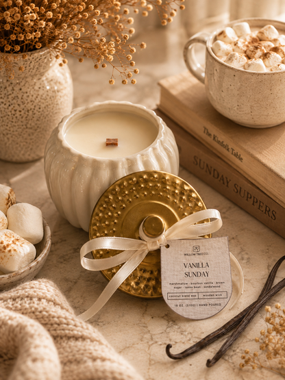 A styled product photo of a scented candle in a ribbed white ceramic jar with its gold lid and cream ribbon tag beside it. The scene includes a mug of marshmallow coffee, books, dried flowers, and a knit blanket on a warm neutral surface.