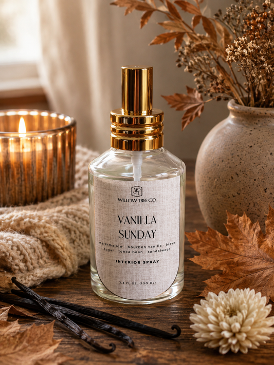 A bottle of Willow Tree Co. Vanilla Sunday interior spray sits on a rustic wooden table surrounded by dried leaves, a lit candle, a knitted blanket, and a ceramic vase with dried botanicals.