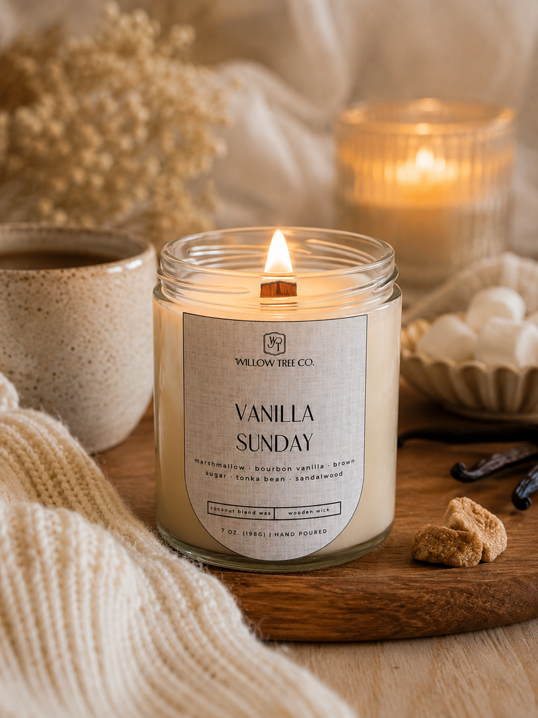 A lit 'Vanilla Sunday' scented candle by Willow Tree Co in a clear glass jar, styled on a wooden board with a mug, marshmallows, and dried flowers in a cozy setting.
