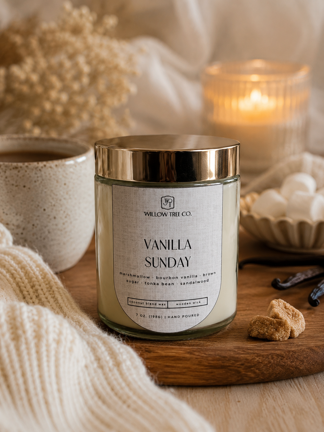 A 'Vanilla Sunday' scented candle by Willow Tree Co. in a clear glass jar with a gold lid, styled on a wooden board with a mug, blanket, and a lit candle in a cozy setting.