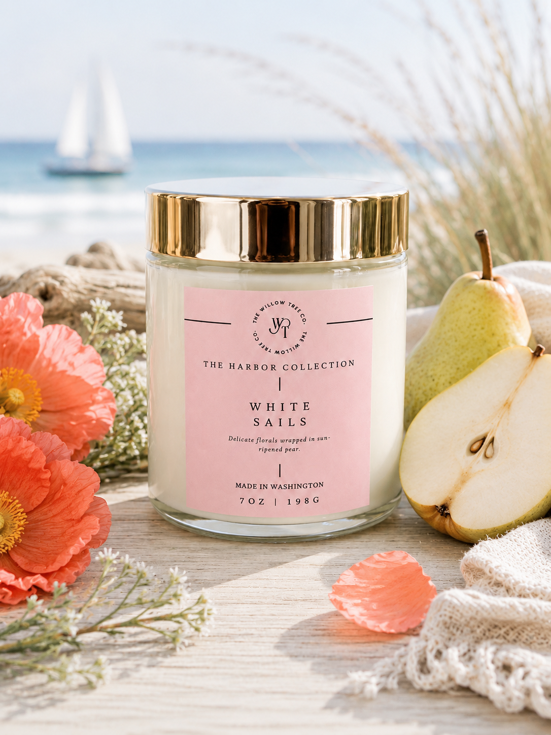 Candle with a pink label on a wooden surface with flowers and a pear, set against a beach backdrop.