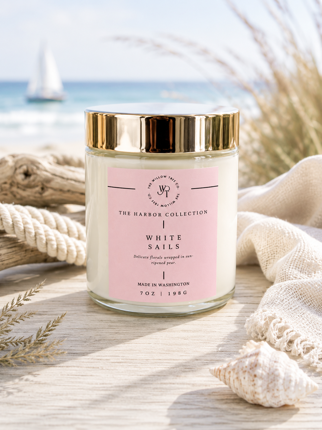 Luxury coastal candle with a soft blush pink label styled beside driftwood, rope, and seashells with an airy ocean backdrop.