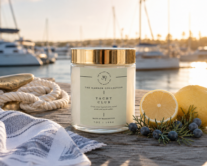 Candle labeled 'Yacht Club' from 'The Harbor Collection' on a wooden surface with lemons and a rope, against a marina backdrop.
