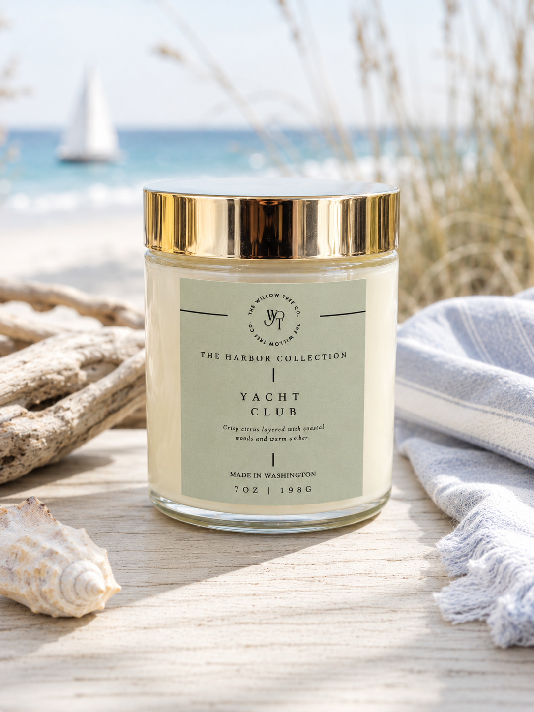 Luxury coastal candle with a pastel sage green label styled beside driftwood and seashells with soft ocean views in the background.
